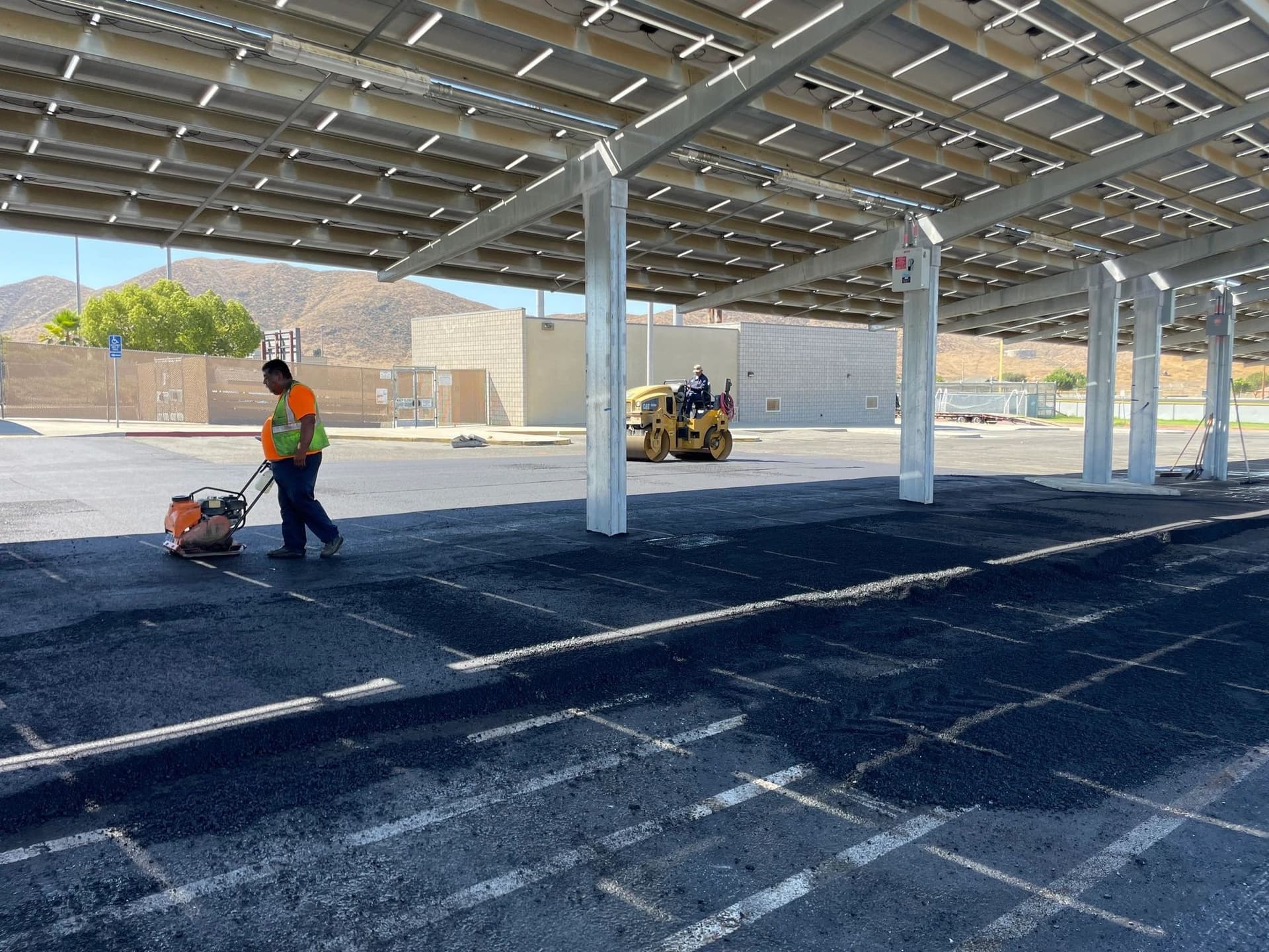 Crew member running a plate compactor over asphalt patches in a striped lot under solar carports