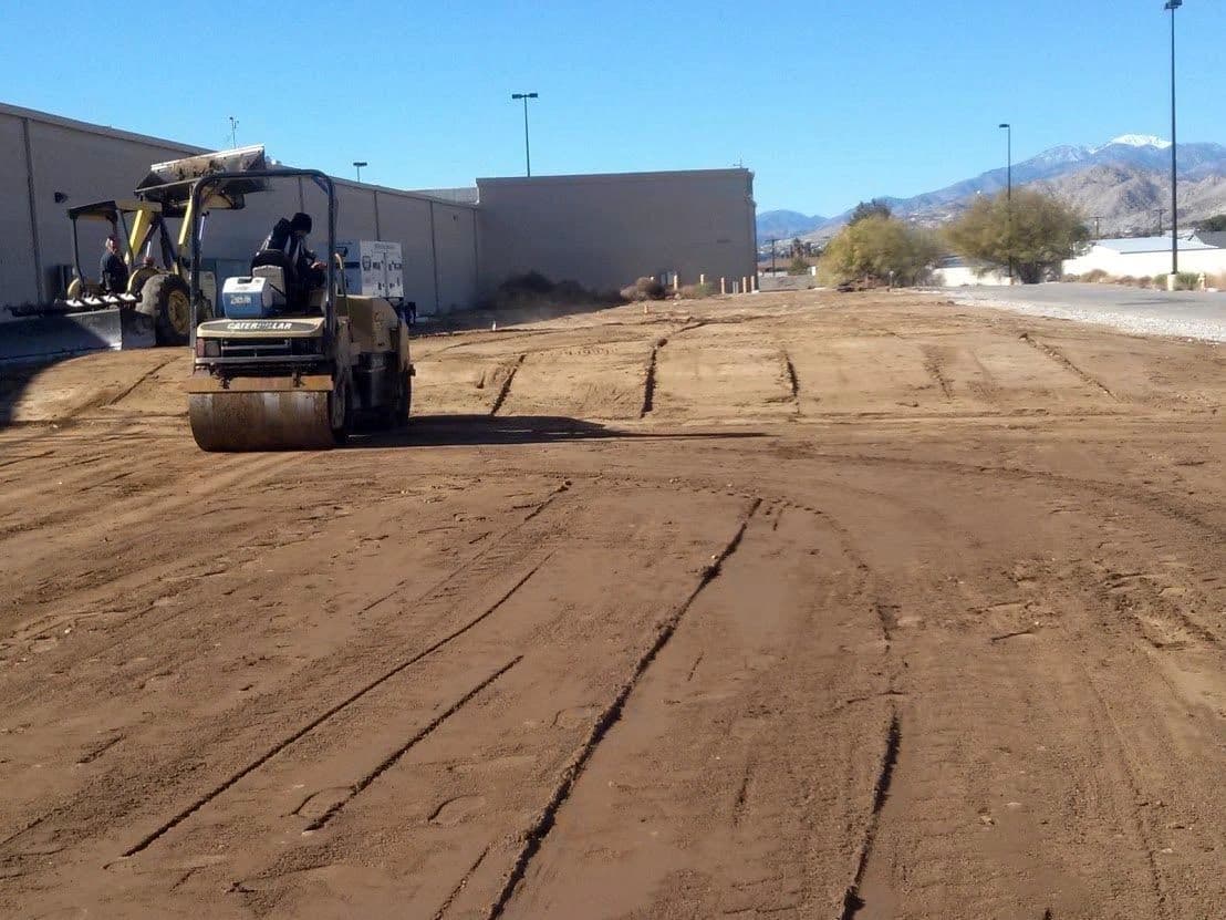 Double-drum roller compacting a graded dirt lot with mountains in the distance