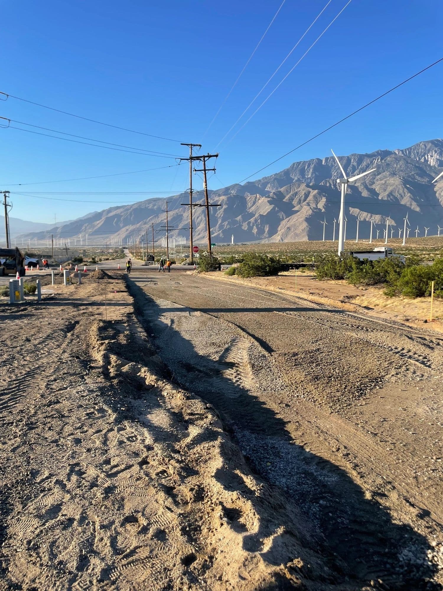 Excavated trench running along a dirt roadside toward distant mountains and wind turbines